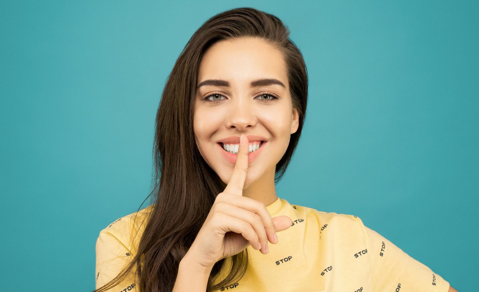 Portrait of a smiling woman making a shushing gesture against a blue background.